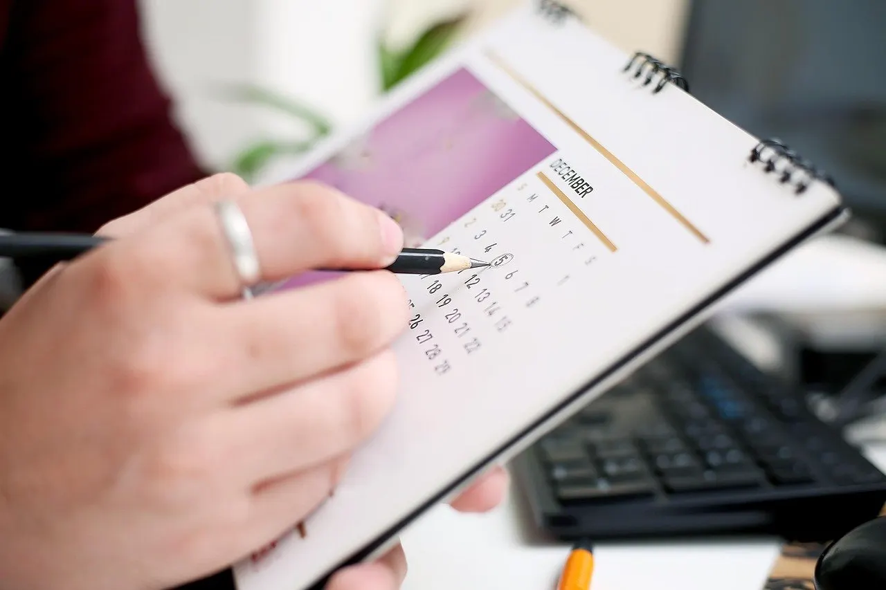 Hand notiert mit Bleistift Termine in einem Spiralblock mit Monatsübersicht, Tastatur im Hintergrund.