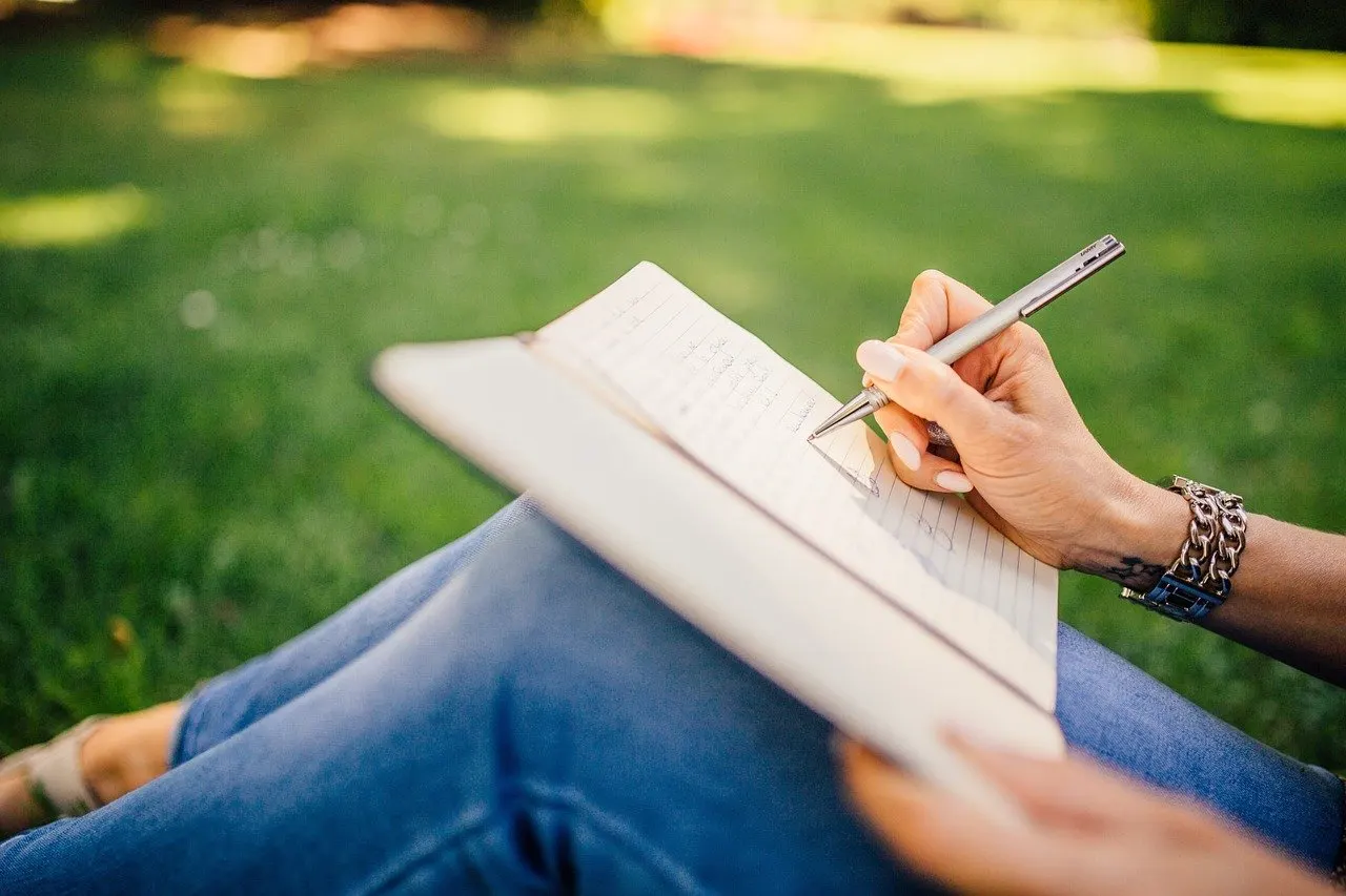 Hand mit Stift schreibt in ein aufgeschlagenes Notizbuch, im Hintergrund grünes Gras, Person sitzt im Freien.