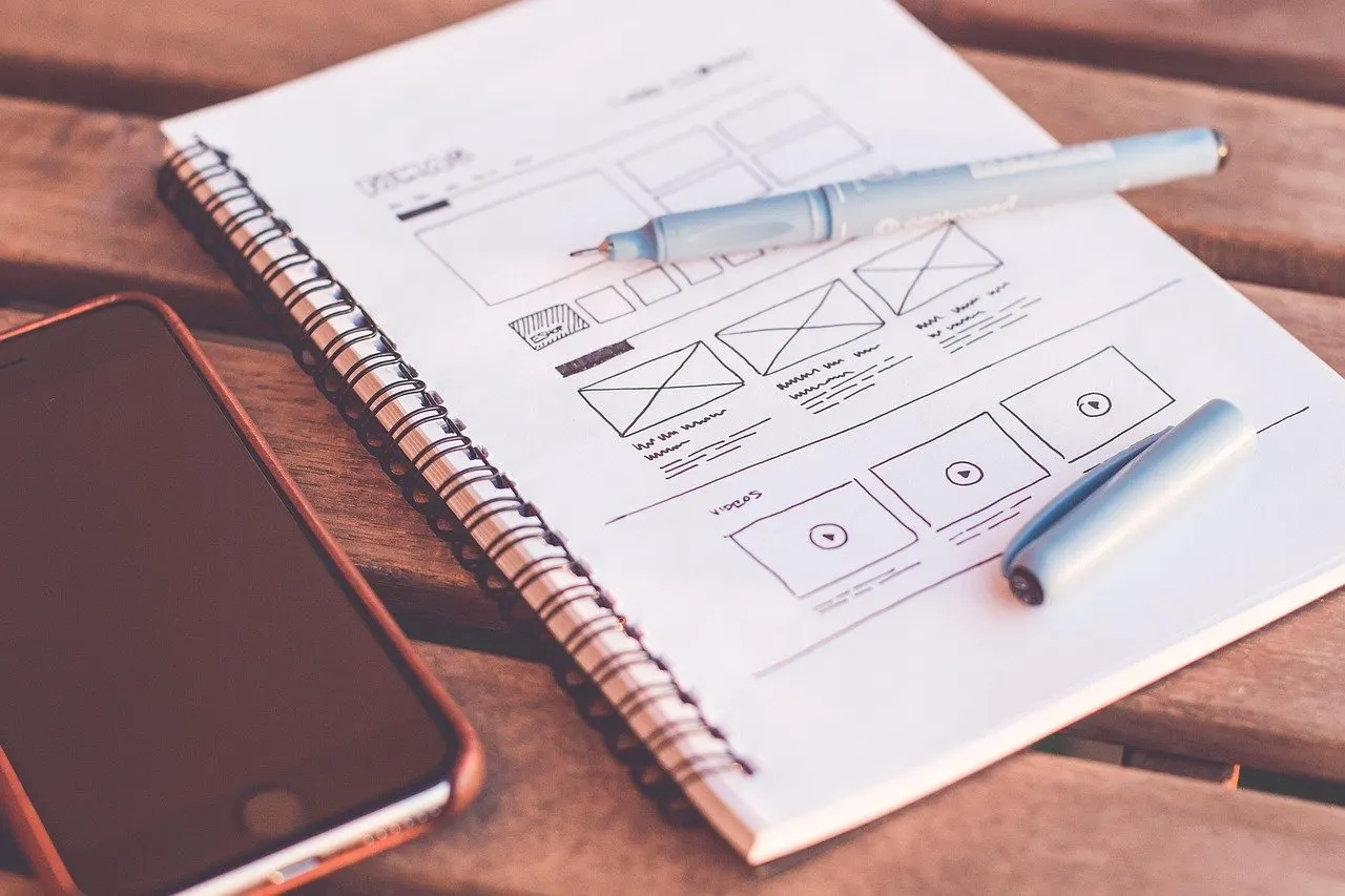 Aufgeschlagenes Notizbuch mit Wireframe-Skizzen, daneben Stift und Smartphone auf einem Holztisch