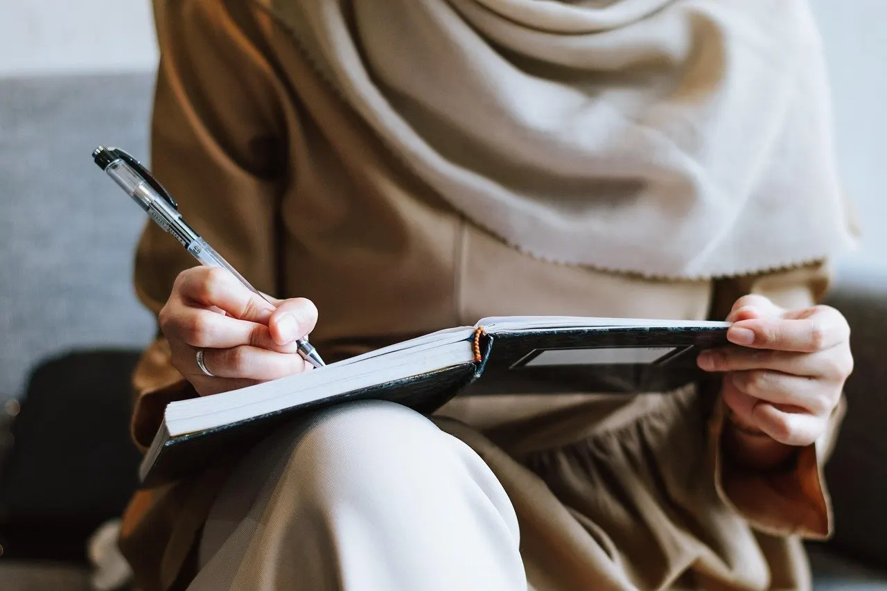 Person mit Schal schreibt mit einem Stift in ein aufgeschlagenes Notizbuch auf dem Schoß.