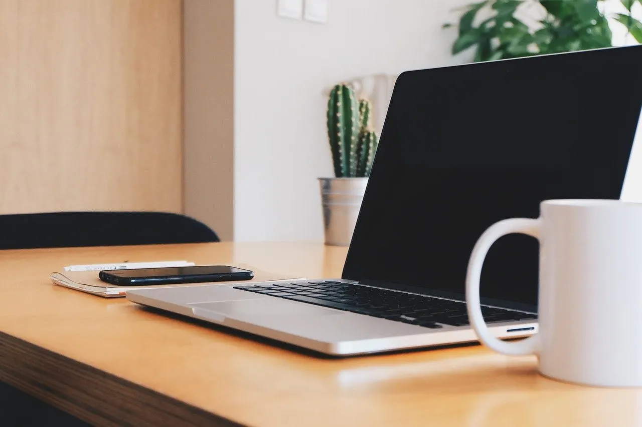 Aufgeklappter Laptop mit schwarzem Bildschirm auf Holztisch, daneben Smartphone und weiße Tasse