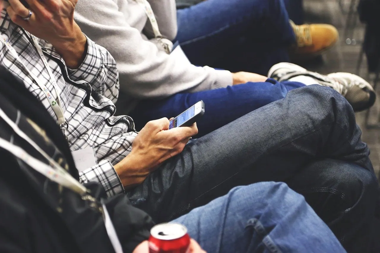 Mehrere Personen sitzen nebeneinander, eine Person hält ein Smartphone, daneben eine Getränkedose.