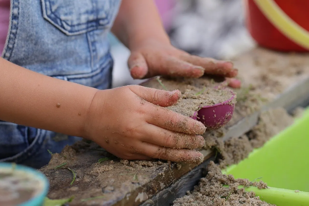 Kinderhände spielen mit Sand in einer Sandkiste, daneben eine pinkfarbene Schaufel.